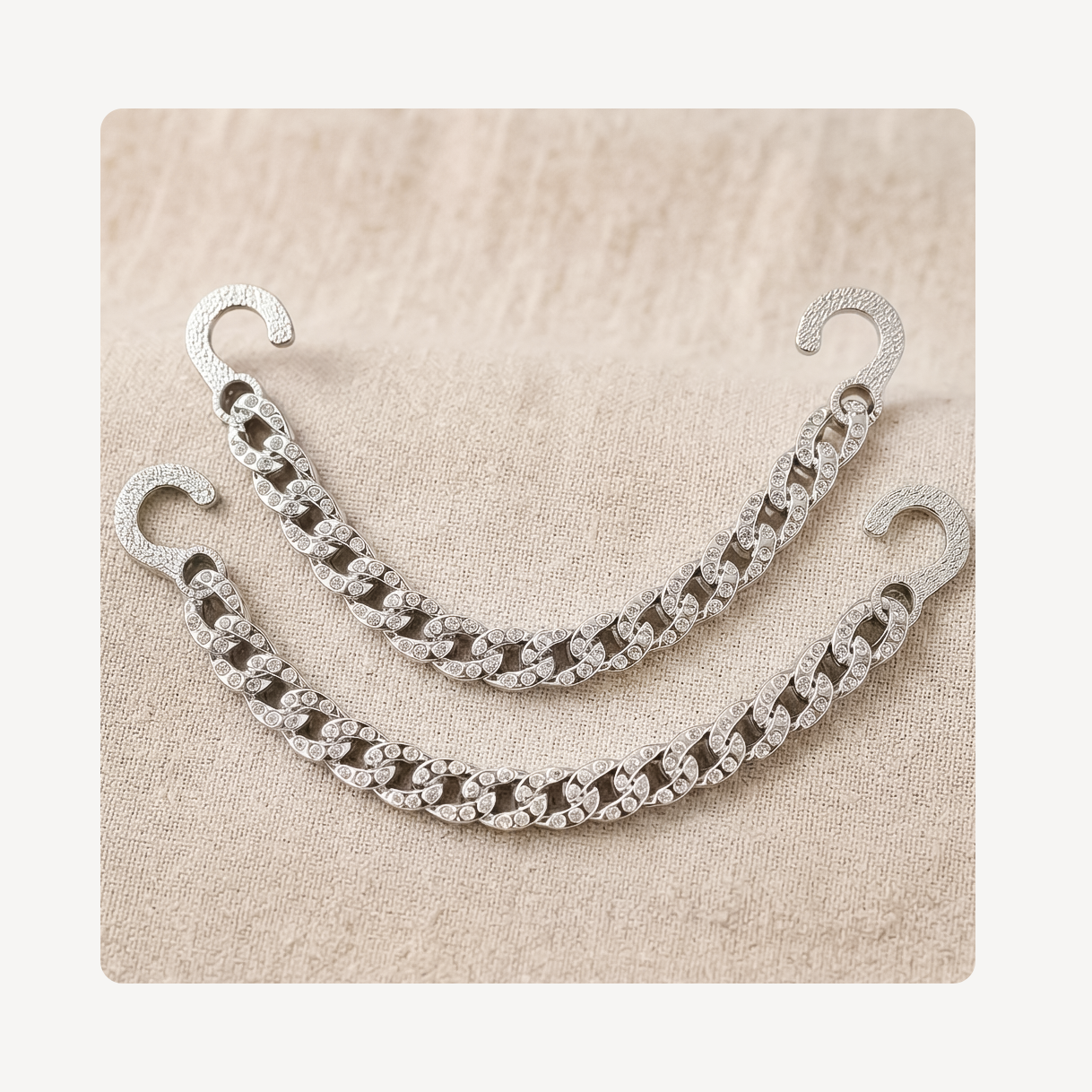 A pair of silver-toned chain link clog chains with hook backs, decorated with clear rhinestones, displayed on a textured beige fabric background.