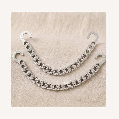 A pair of silver-toned chain link clog chains with hook backs, decorated with clear rhinestones, displayed on a textured beige fabric background.