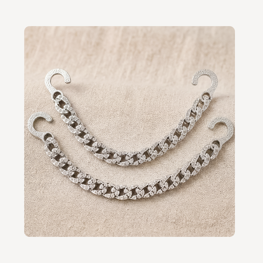 A pair of silver-toned chain link clog chains with hook backs, decorated with clear rhinestones, displayed on a textured beige fabric background.