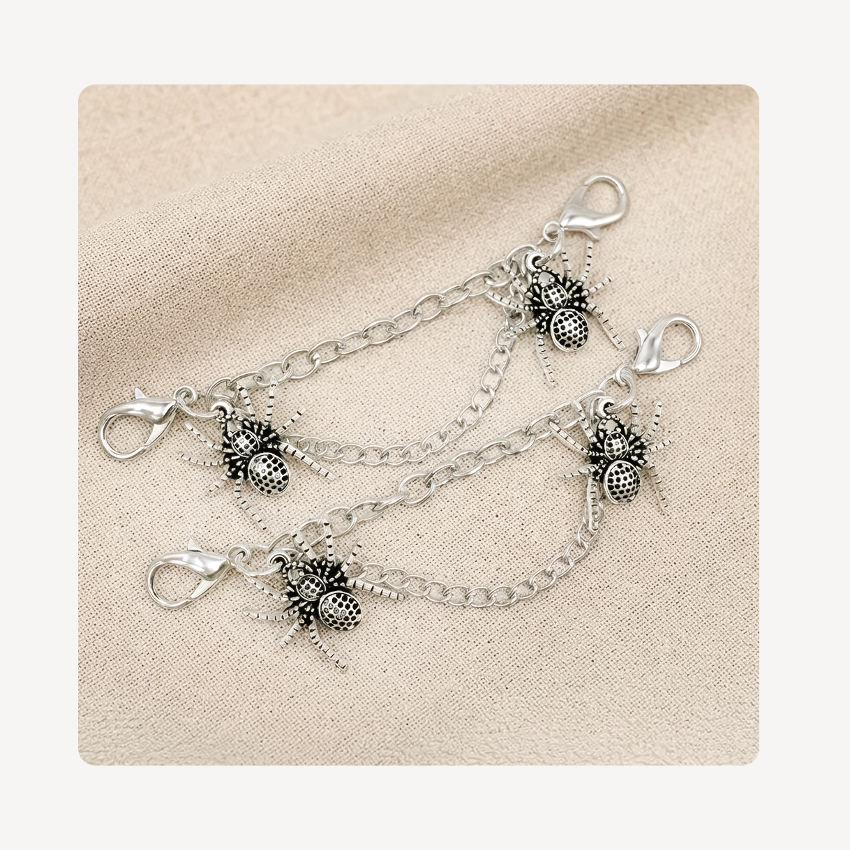 Two silver shoe chains with spider charms, displayed on a textured beige fabric background.