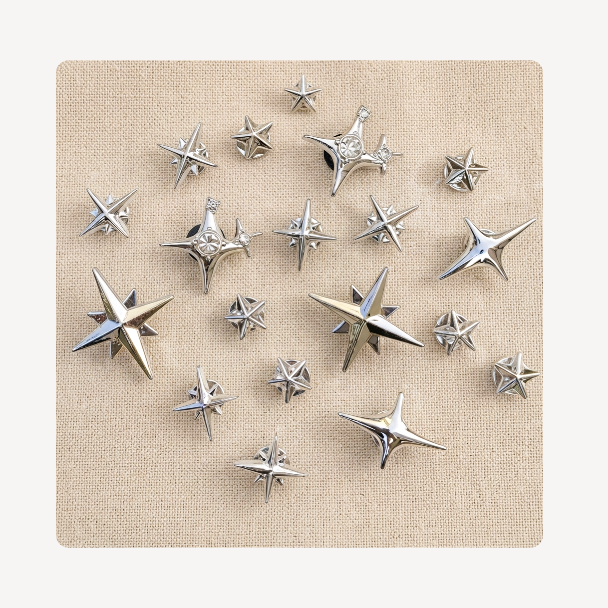 A set of metallic silver star-shaped clog charms arranged on a square beige fabric backdrop against a light off-white background.