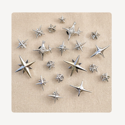 A set of metallic silver star-shaped clog charms arranged on a square beige fabric backdrop against a light off-white background.