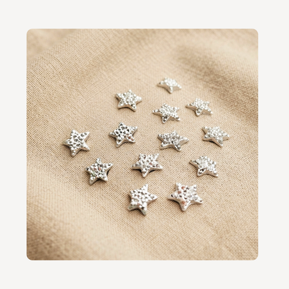 A group of small silver star-shaped clog charms with a textured surface, arranged on a beige linen fabric background.