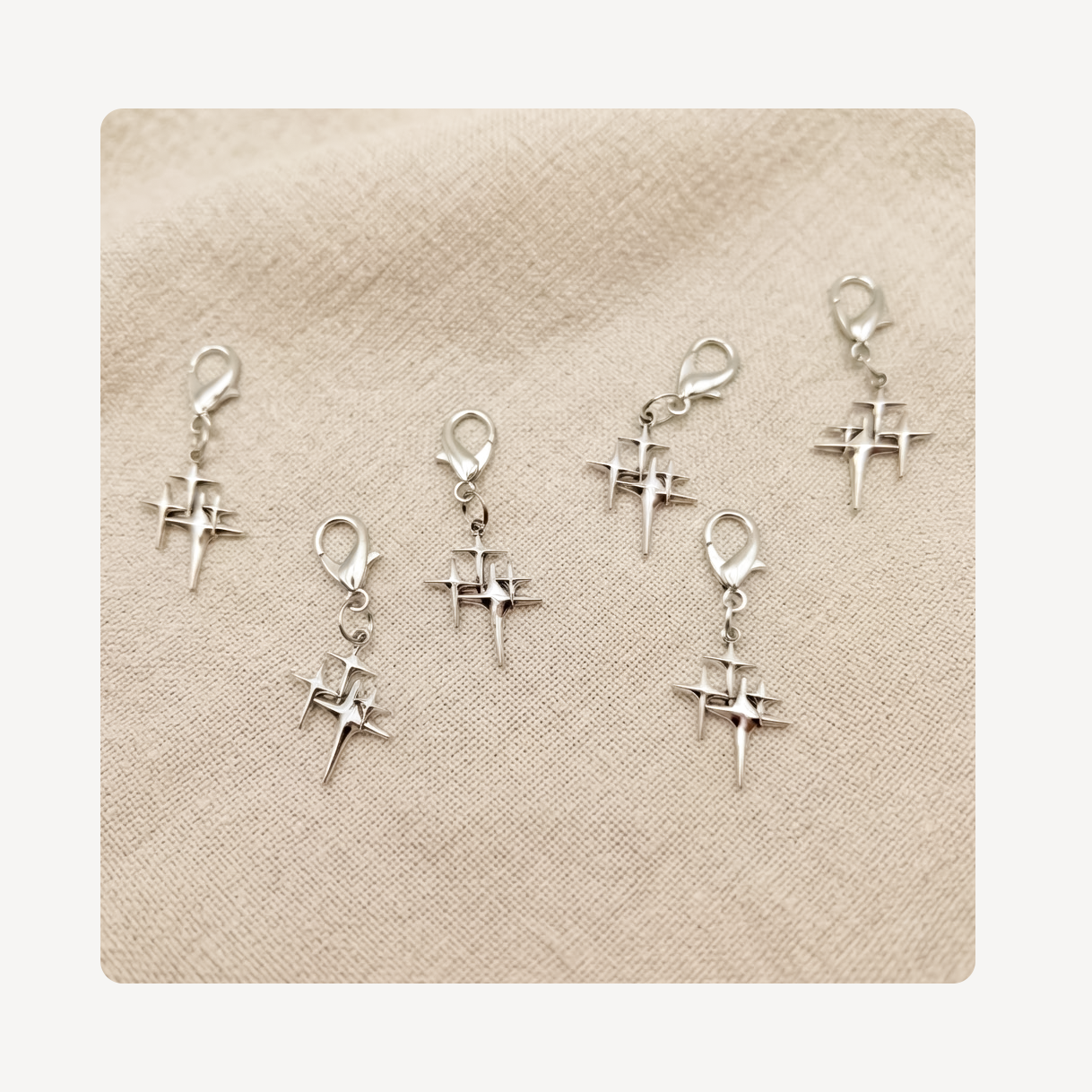 Six silver star shoe charms with lobster clasp closures, arranged on a textured beige fabric background.