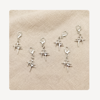 Six silver star shoe charms with lobster clasp closures, arranged on a textured beige fabric background.
