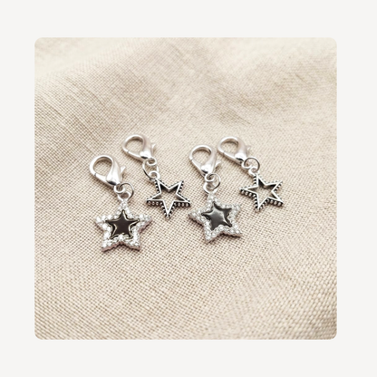 Four silver-toned star-shaped shoe charm pendants with lobster clasps, arranged on a textured beige fabric background.
