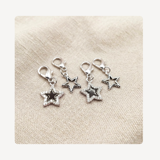 Four silver-toned star-shaped shoe charm pendants with lobster clasps, arranged on a textured beige fabric background.