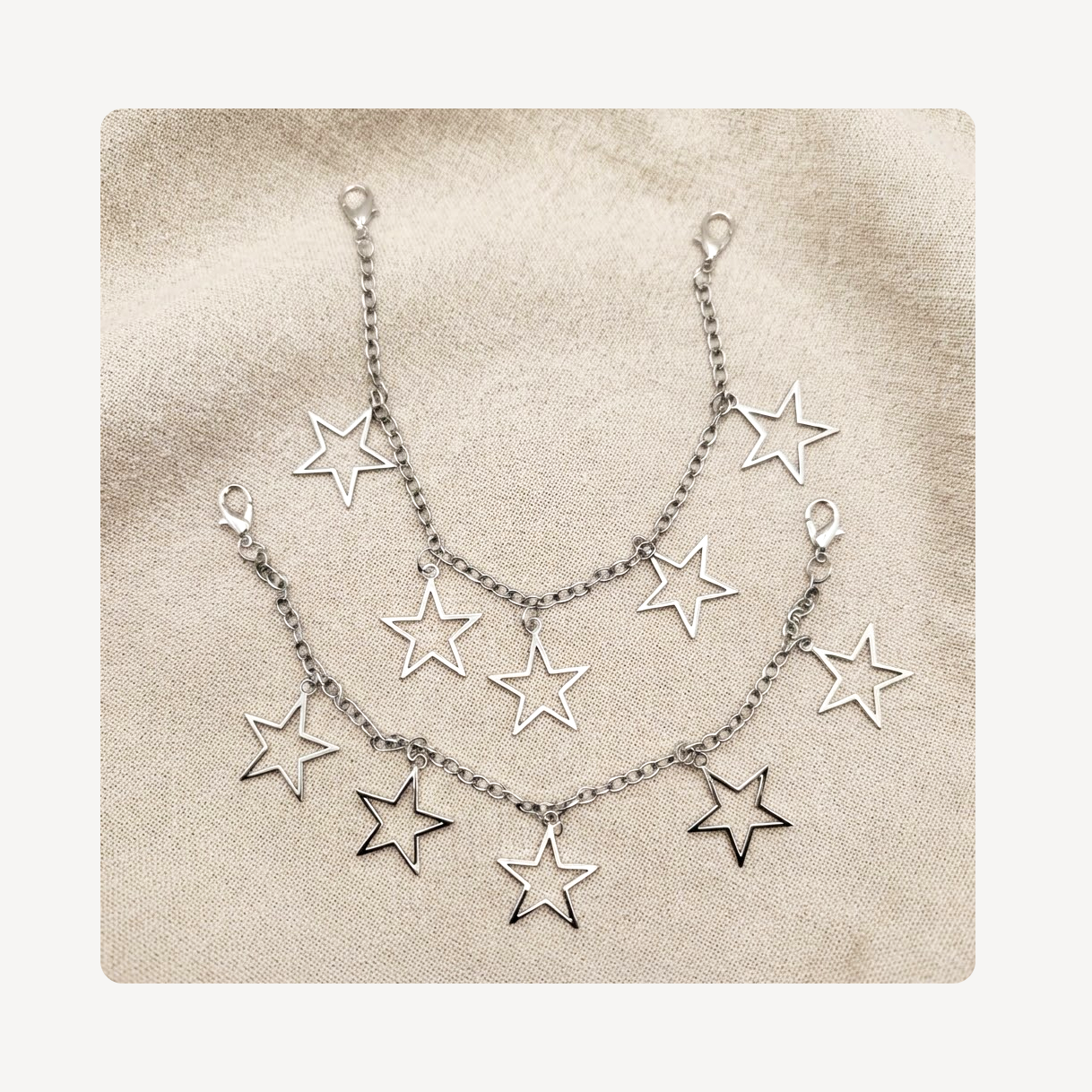 A silver-toned double-layered shoe chain featuring multiple dangling open star charms, displayed on a textured beige fabric background.