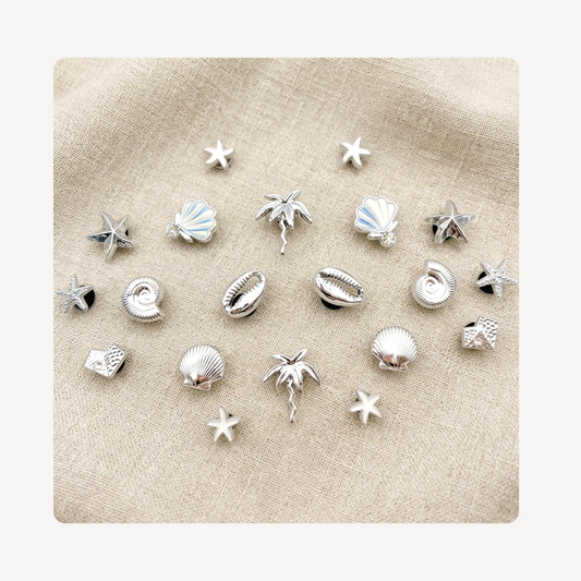 A set of small silver-toned shell and starfish clog charms arranged on a beige textured fabric background.