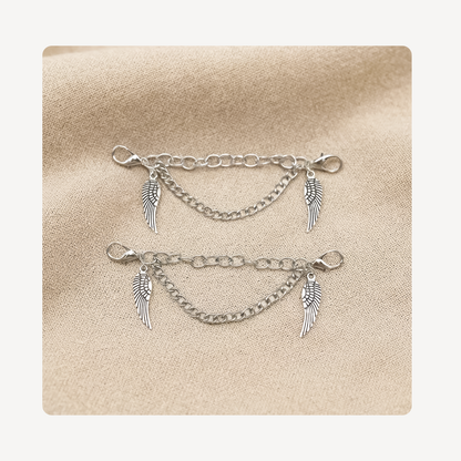 A pair of silver angel wing shoe chains with lobster clasp closures, displayed on a textured beige fabric background.