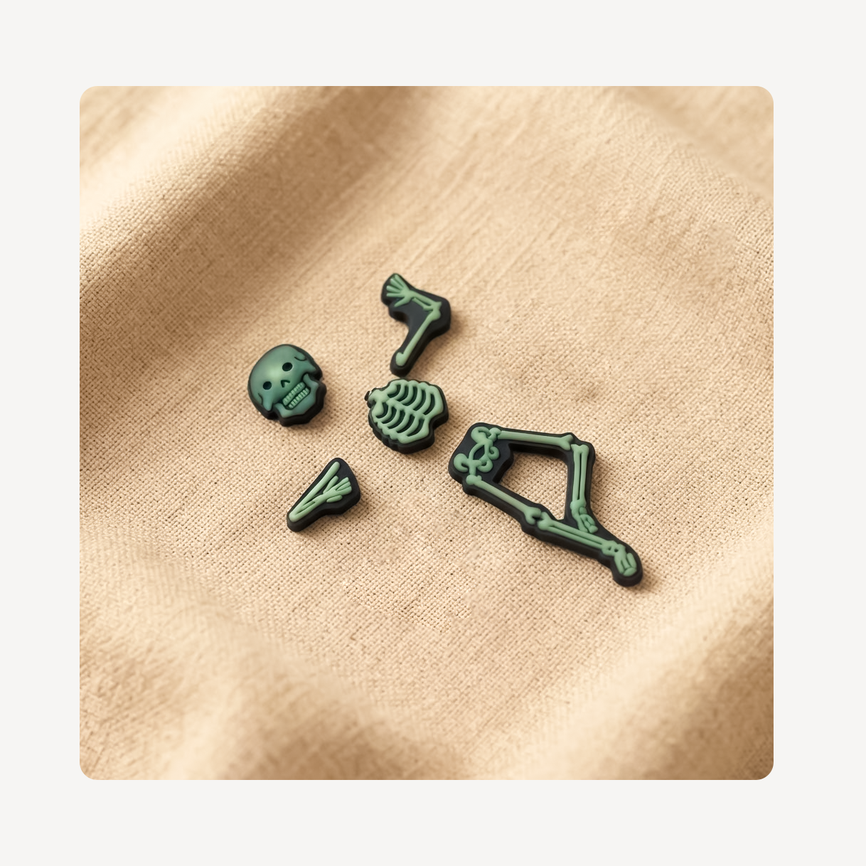 Five small dark green skeleton-themed clog charms arranged on a beige fabric background.
