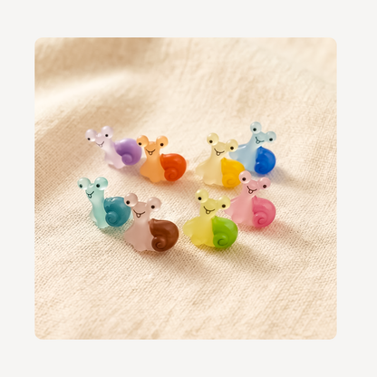 Six small pastel snail-shaped clog charms arranged on a soft beige fabric background. The set includes colorful snail-like figures in purple, orange, yellow, blue, teal, pink, brown, and green.