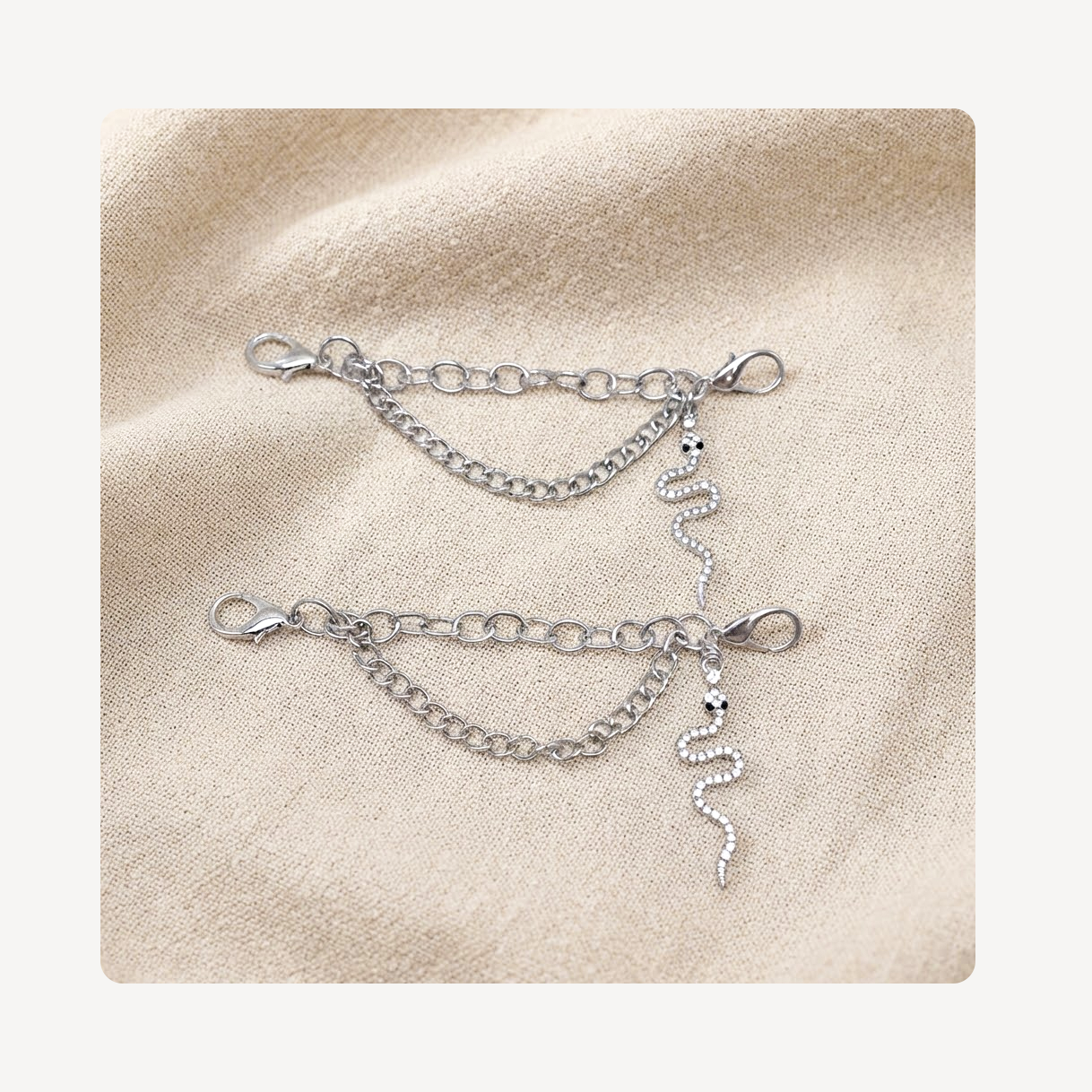 A pair of silver shoe chains with lobster clasp closures, displayed on a textured beige fabric background.