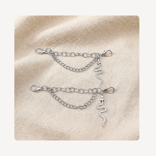 A pair of silver shoe chains with lobster clasp closures, displayed on a textured beige fabric background.