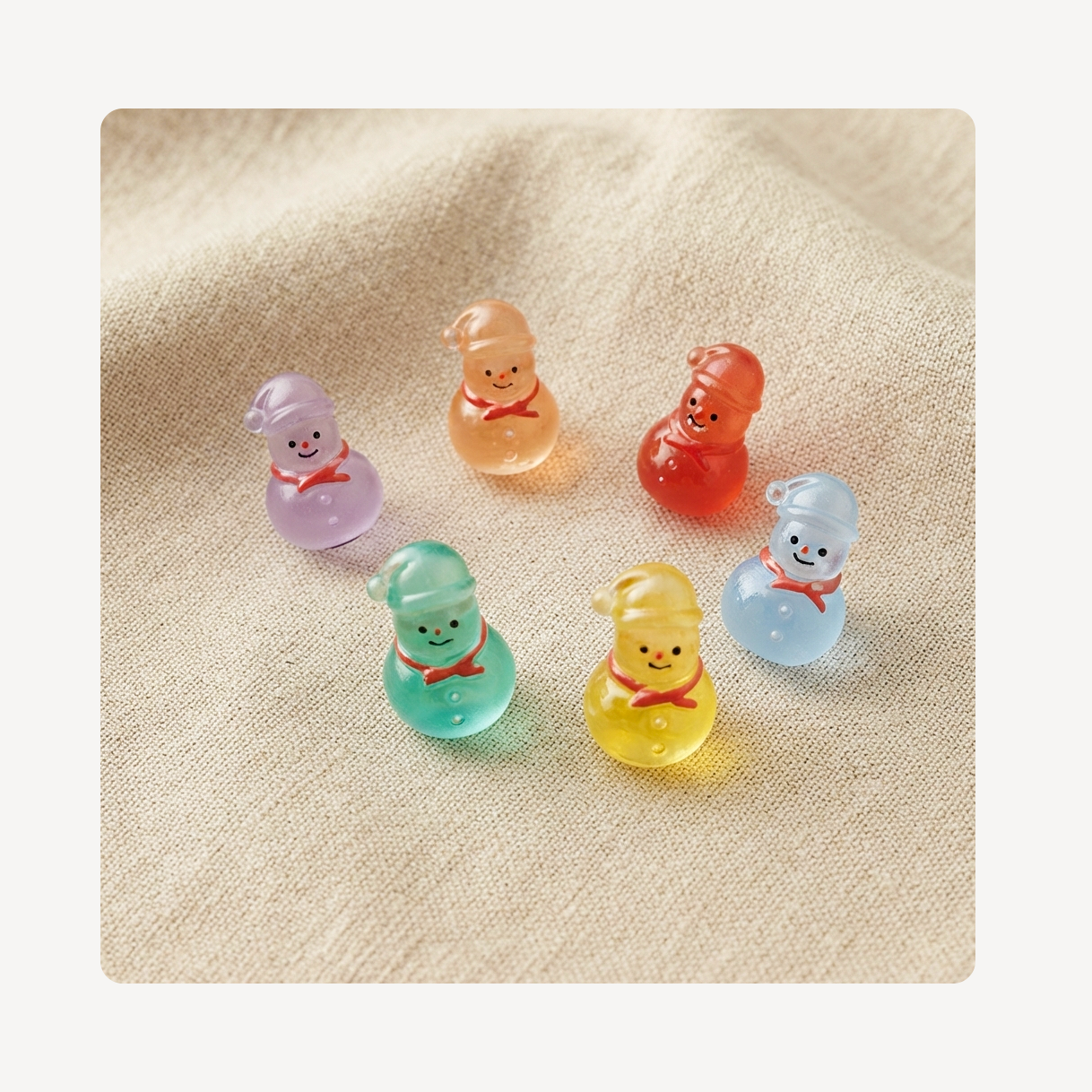 Six small translucent snowman clog charms in pastel colors, arranged on a textured beige fabric background.