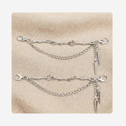 A pair of silver shoe chains with spiked dangling charms, displayed on a textured beige fabric background.