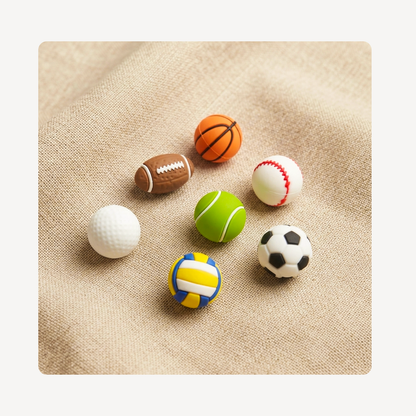 Seven miniature sports-themed clog charms shaped like a basketball, football, baseball, golf ball, tennis ball, soccer ball, and volleyball, arranged on a textured beige linen background.