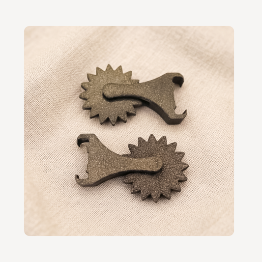 Two dark cowboy spur clog charms with hook-like ends, displayed on a textured beige fabric background.