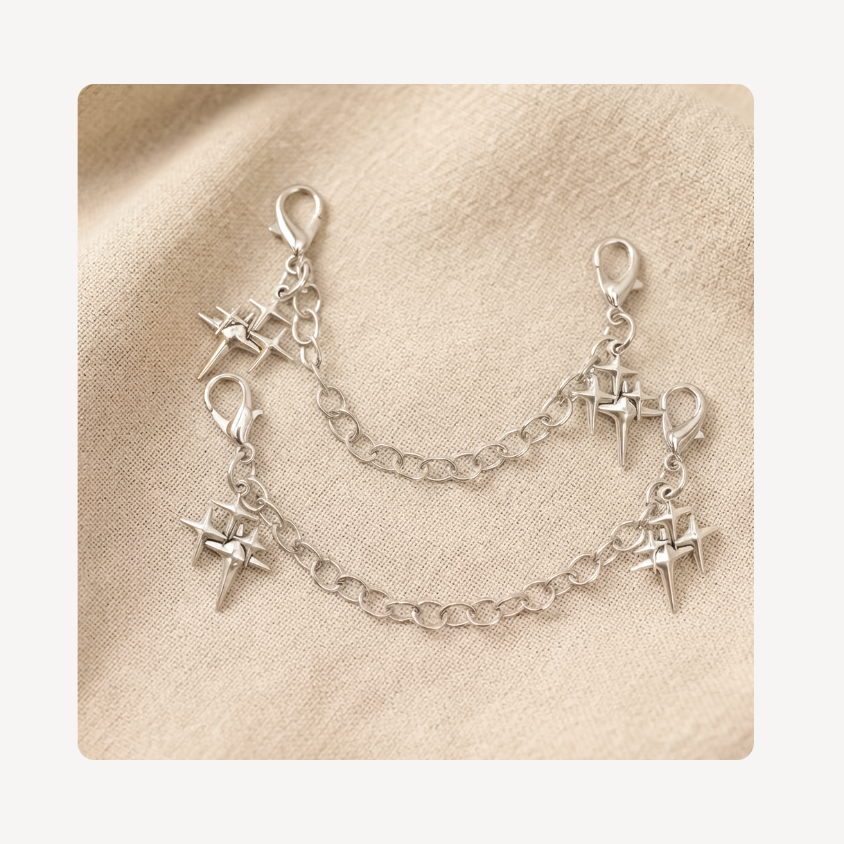 A pair of silver shoe chains with lobster clasp ends and dangling starburst charms, displayed on a textured beige fabric background.