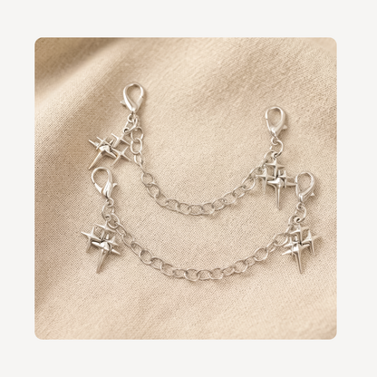 A pair of silver shoe chains with lobster clasp ends and dangling starburst charms, displayed on a textured beige fabric background.