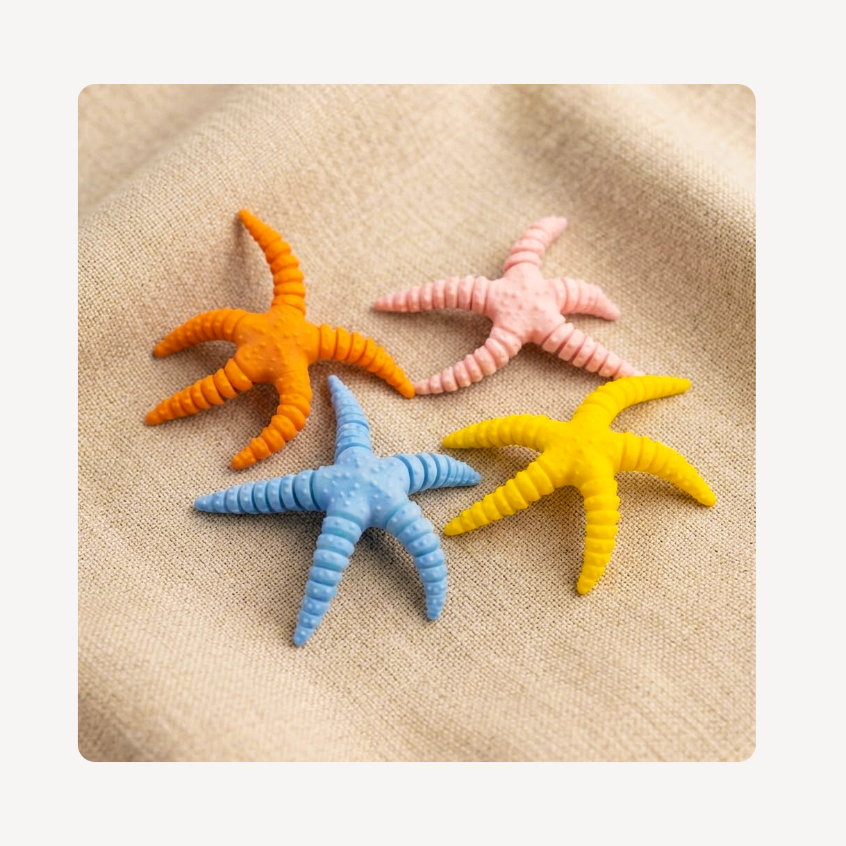 Four textured starfish-shaped clog charms in orange, pink, blue, and yellow, arranged on a beige fabric background.