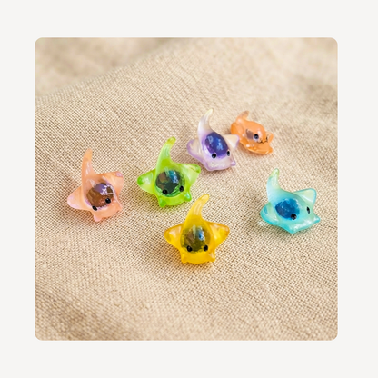 Six small translucent stringray clog charms in pastel colors including peach, green, lavender, orange, and blue, arranged on a beige textured fabric background.