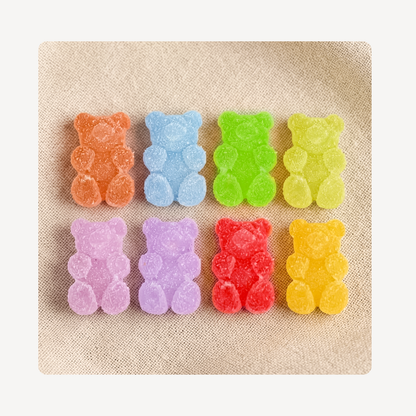 Eight colorful gummy bear candy clog charms arranged in two rows on a textured beige fabric background.