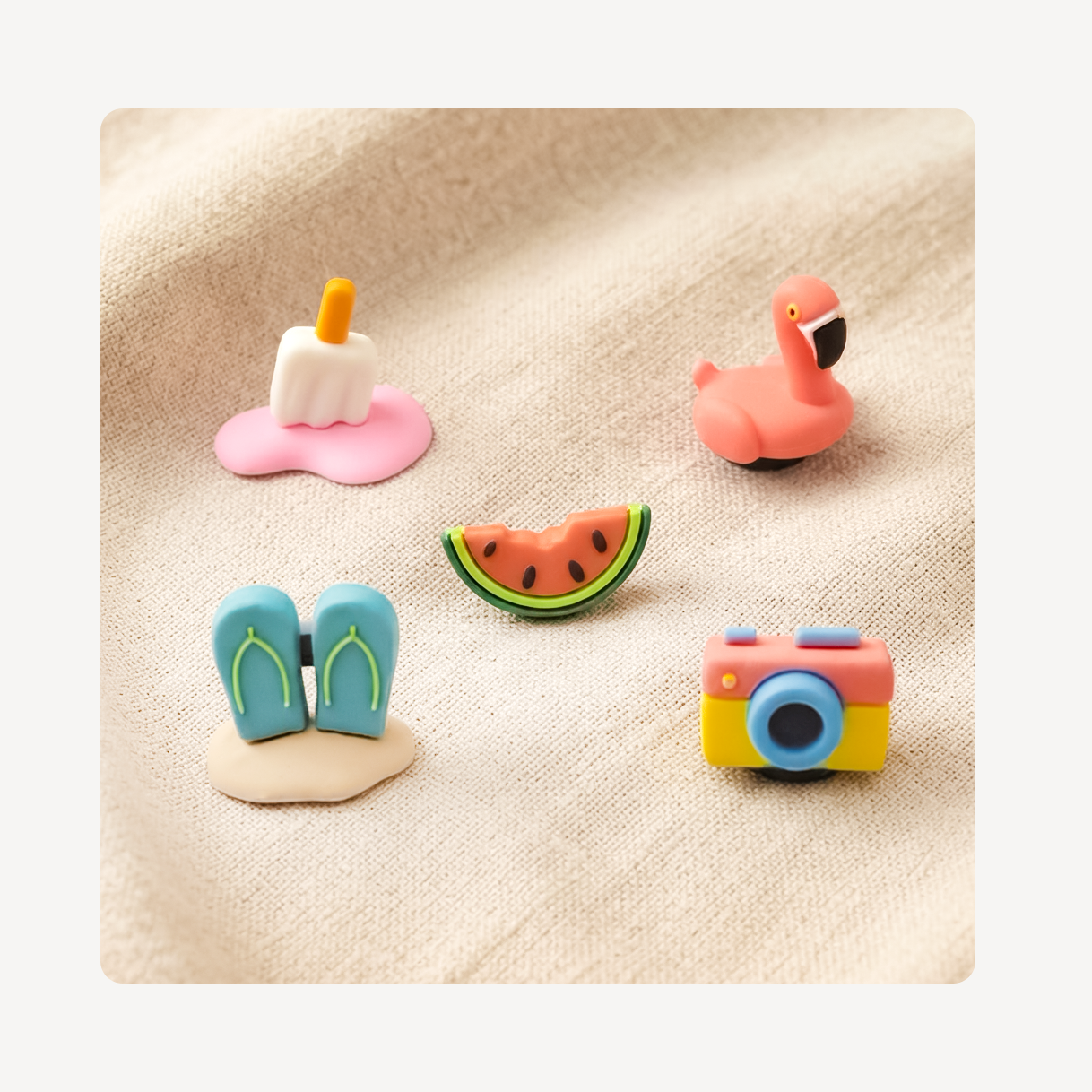Five small colorful summer-themed clog charms arranged on a beige textured fabric background: a pink flamingo, blue flip-flops, a watermelon slice, and a pink-and-yellow camera.