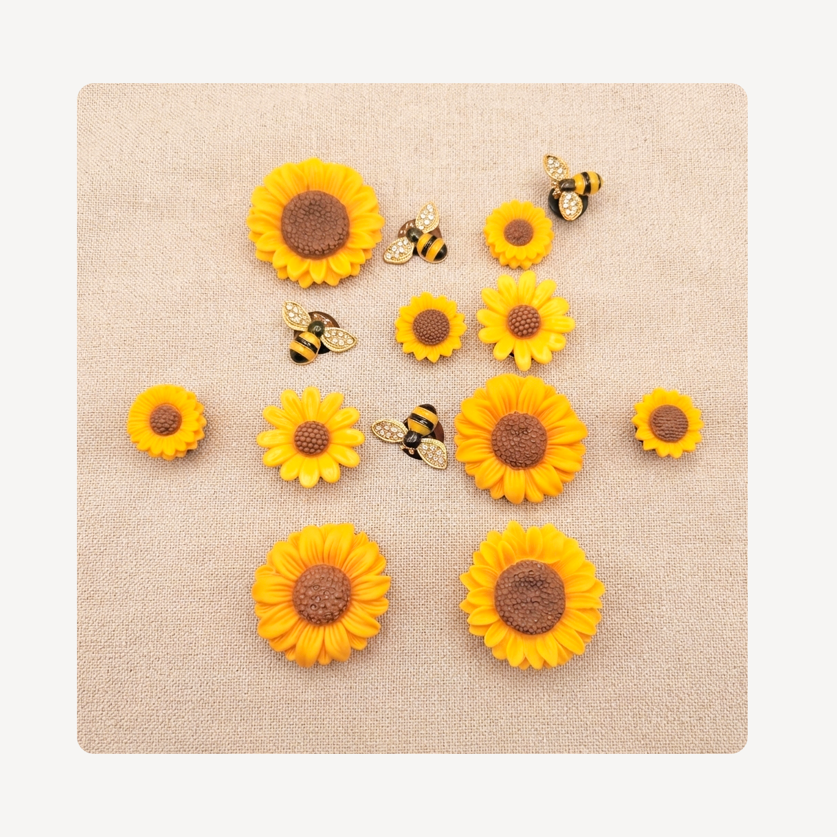 A flat-lay arrangement of yellow sunflower-like clog charms and three bumblebees on a square beige textured mat against a white background.