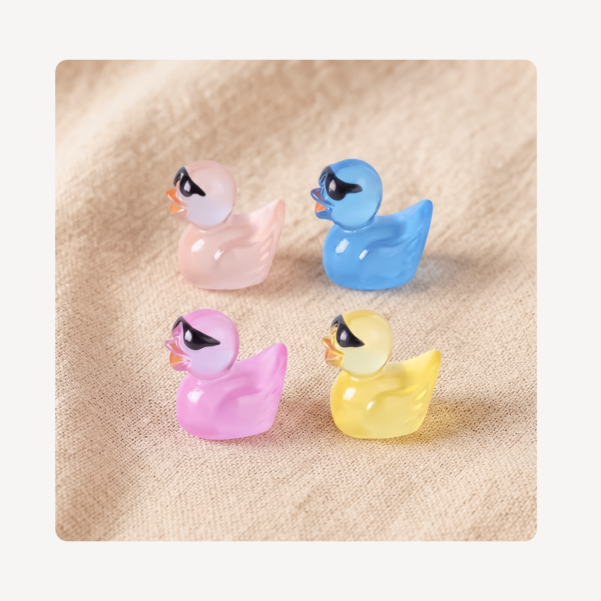 Four small glossy ducky clog charms arranged in a 2x2 grid on a beige textured fabric background. The ducks are translucent pastel pink, blue, purple, and yellow, each with a black-and-white stylized face and orange beaks.