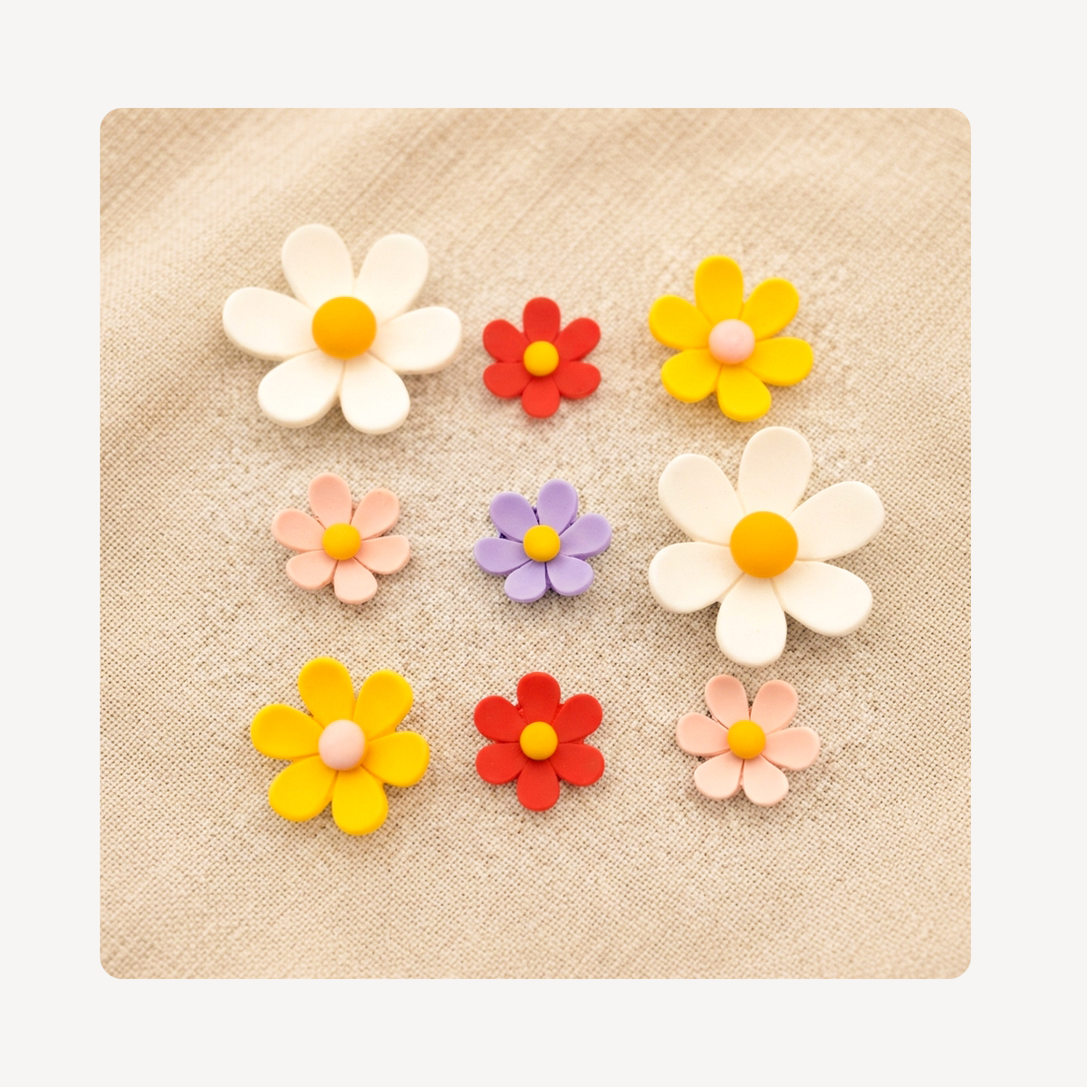 Nine colorful flower-shaped clog charms arranged on a beige textured fabric background. The flowers include white, yellow, red, peach, and lavender colors, with some featuring orange centers and others pink centers.
