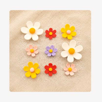Nine colorful flower-shaped clog charms arranged on a beige textured fabric background. The flowers include white, yellow, red, peach, and lavender colors, with some featuring orange centers and others pink centers.