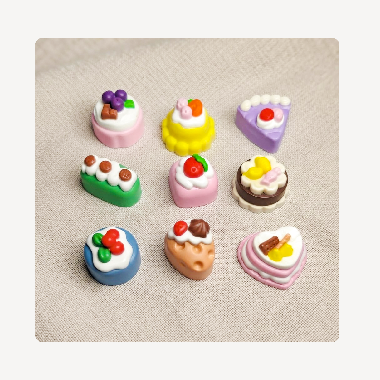 Nine miniature cake clog charms arranged in a 3x3 grid on a textured beige fabric background.