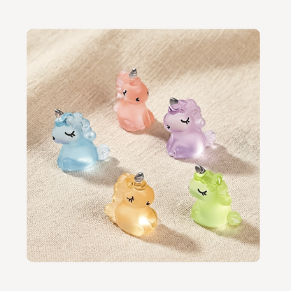 Five small translucent unicorn clog charms in pastel blue, peach, lavender, yellow, and green, arranged on a textured beige fabric background.