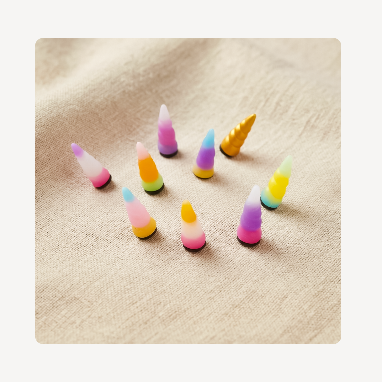 Nine small unicorn horn-shaped clog charms in assorted pastel and bright colors, arranged on a beige textured fabric background.