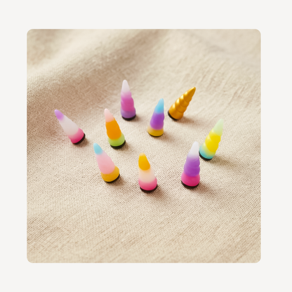 Nine small unicorn horn-shaped clog charms in assorted pastel and bright colors, arranged on a beige textured fabric background.