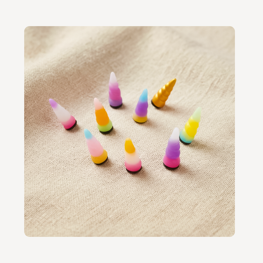 Nine small unicorn horn-shaped clog charms in assorted pastel and bright colors, arranged on a beige textured fabric background.