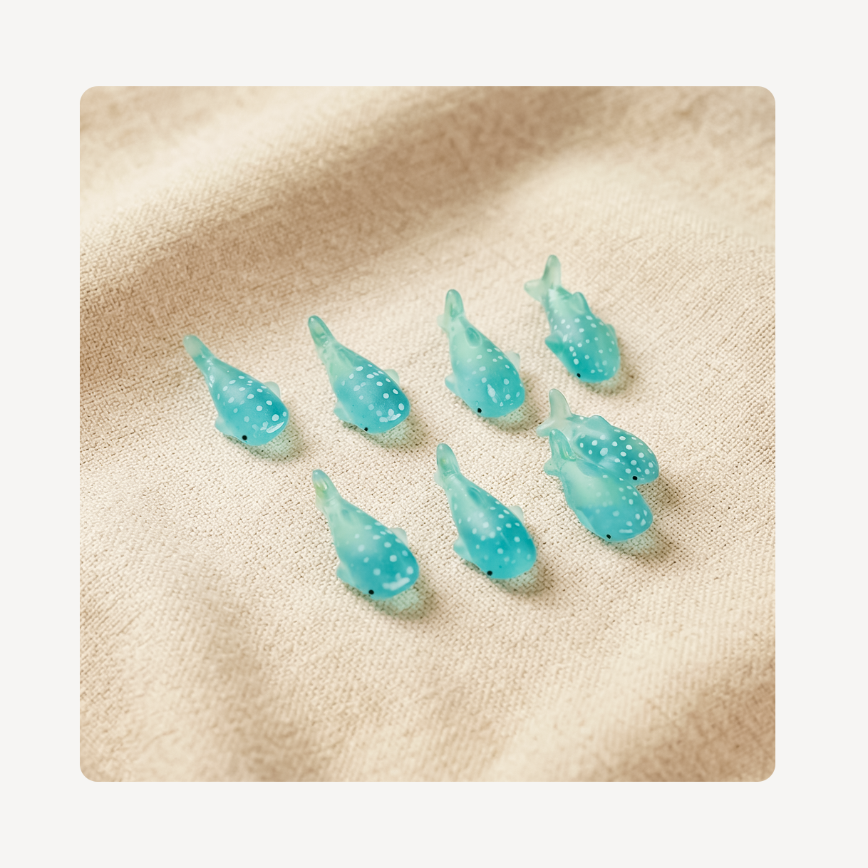Eight small translucent turquoise whale-shaped clog charms with white speckles arranged on a beige textured fabric background.