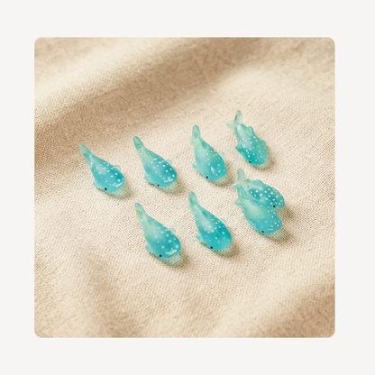 Eight small translucent turquoise whale-shaped clog charms with white speckles arranged on a beige textured fabric background.