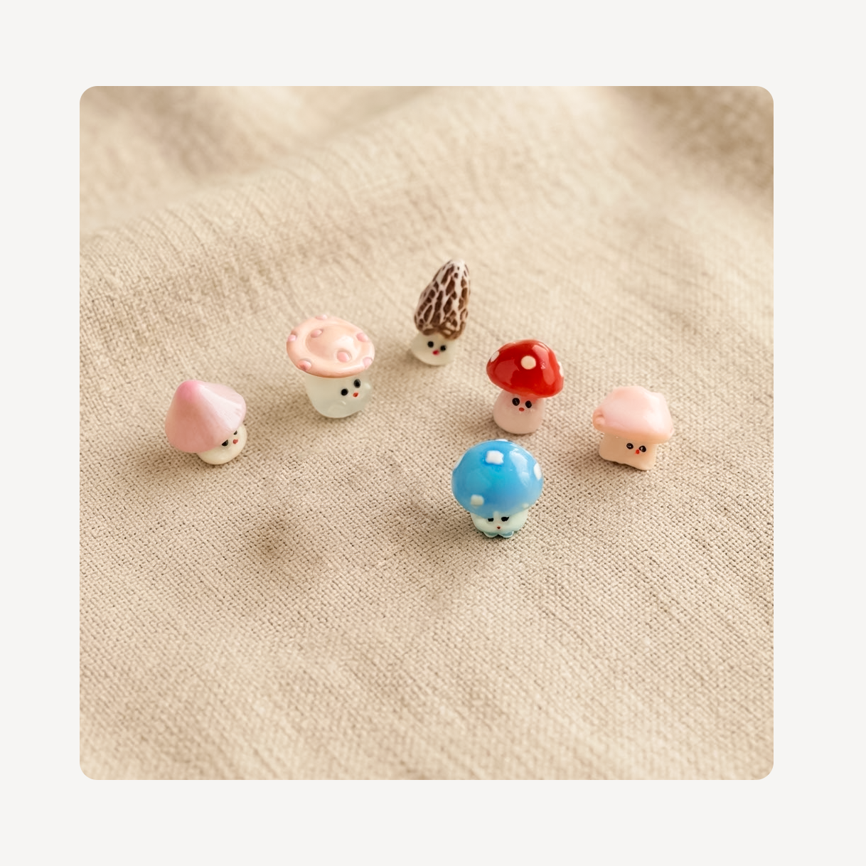 Six small mushroom-shaped clog charms with cute cartoon faces are arranged on a beige linen fabric background.