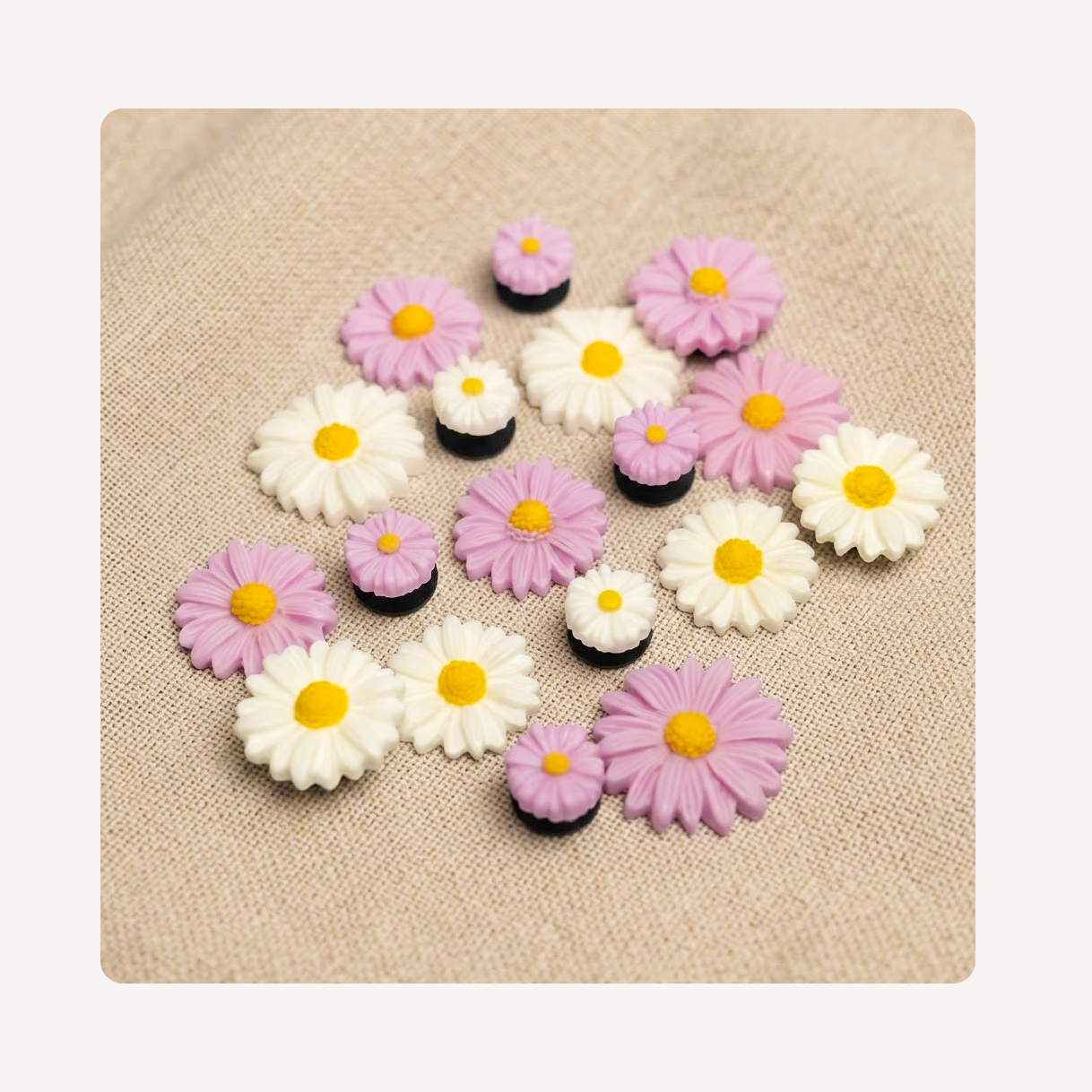 A set of small daisy-shaped clog charms in white, light purple, and black, arranged on a beige textured fabric background.