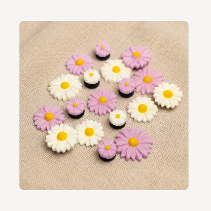 A set of small daisy-shaped clog charms in white, light purple, and black, arranged on a beige textured fabric background.