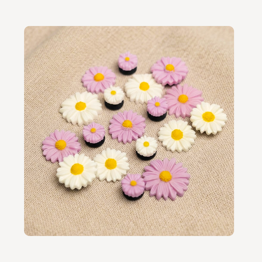 A set of small daisy-shaped clog charms in white, light purple, and black, arranged on a beige textured fabric background.