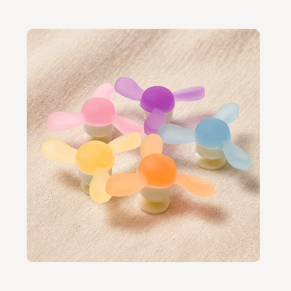 Five small windmill clog charms in pastel colors including pink, purple, blue, yellow, and orange, arranged on a textured beige fabric background.