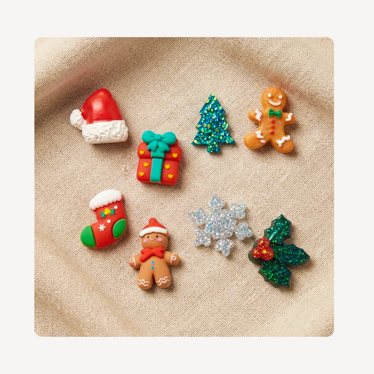 Eight small Christmas-themed decorative clog charms arranged on a beige fabric background: a red Santa hat, a red gift box with a green bow, a glittery green tree, a gingerbread man, a red stocking with green and white trim, a silver glitter snowflake, and a green holly leaf with red berries.