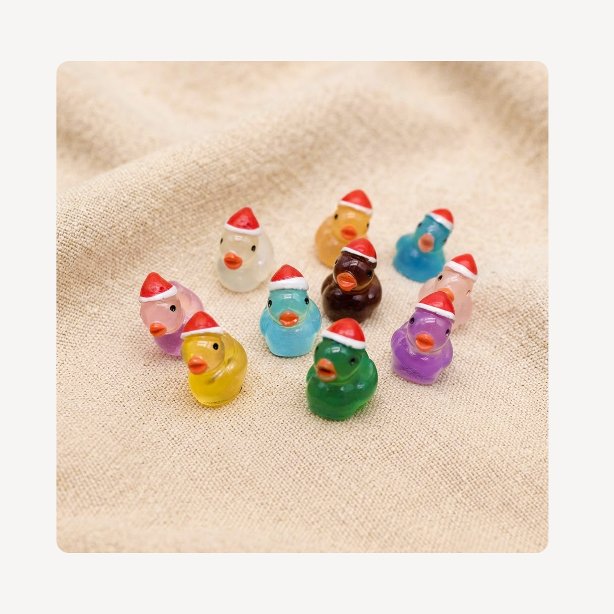 A group of small translucent duck clog charms in assorted colors, each wearing a red Santa hat, arranged on a beige textured fabric background.