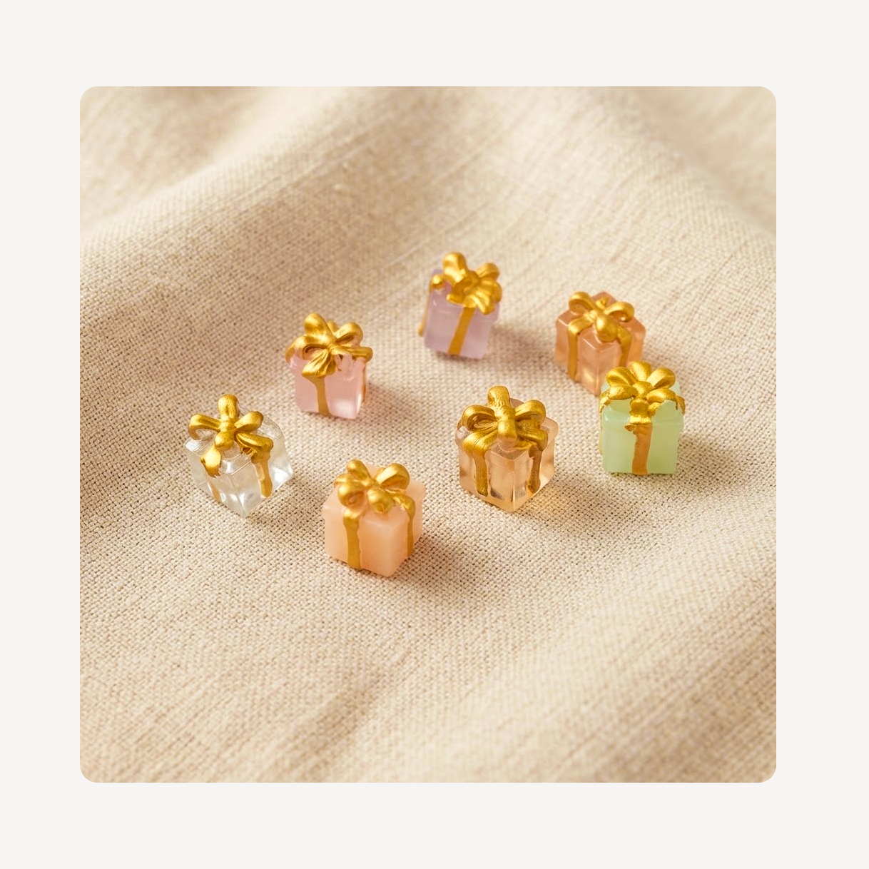 Six small gift box clog charms with gold ribbon bows arranged on a beige textured fabric background.