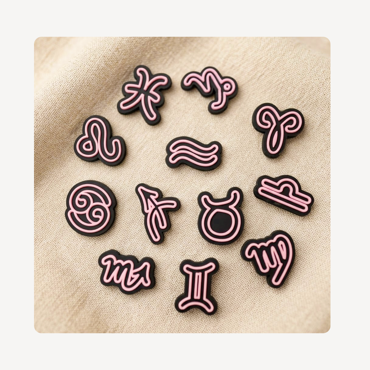 Twelve astrological zodiac sign clog charms arranged in a circle on a beige textured fabric background. Each charm features a pink neon-style outline of a zodiac symbol with a black fill.