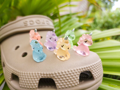 Beige Crocs decorated with glow-in-the-dark unicorn charms in various pastel colors, including purple, green, pink, orange, yellow, and blue, placed on grass. The charms add a whimsical and magical touch to the shoes.