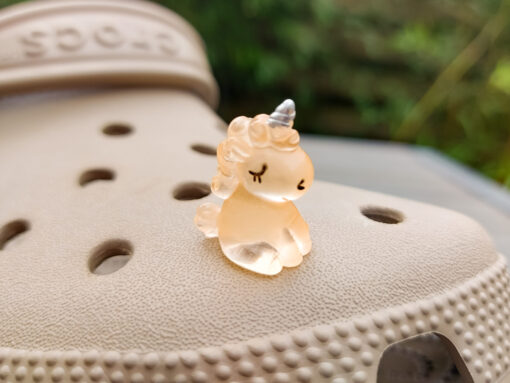 Beige Crocs decorated with glow-in-the-dark unicorn charms in various pastel colors, including purple, green, pink, orange, yellow, and blue, placed on grass. The charms add a whimsical and magical touch to the shoes.